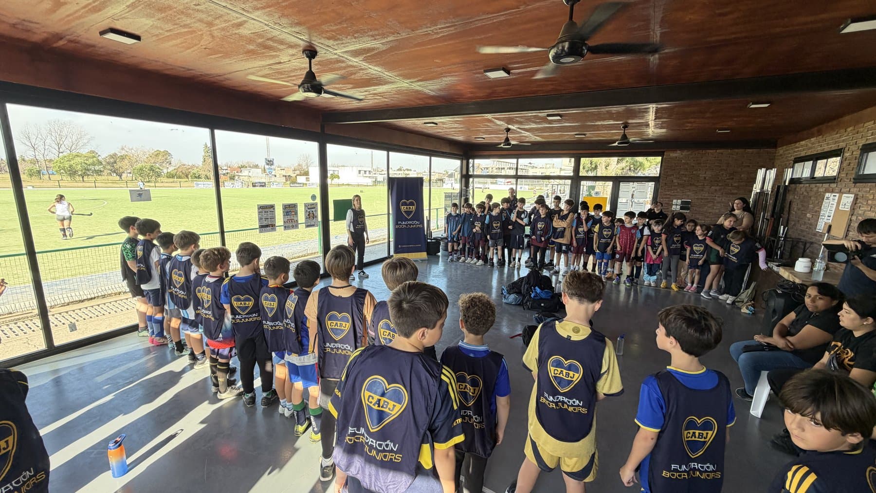 Chicos y chicas con pecheras de la Fundación Boca Juniors formados en el salón de un club de barrio, escuchando la charla
