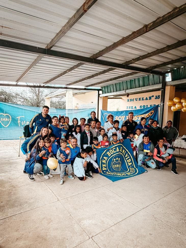 Chicos, chicas y adultos posando en el patio de la Escuela N.° 837 Isla Soledad, con la bandera de la Peña Boca de mi Vida de Santiago del Estero