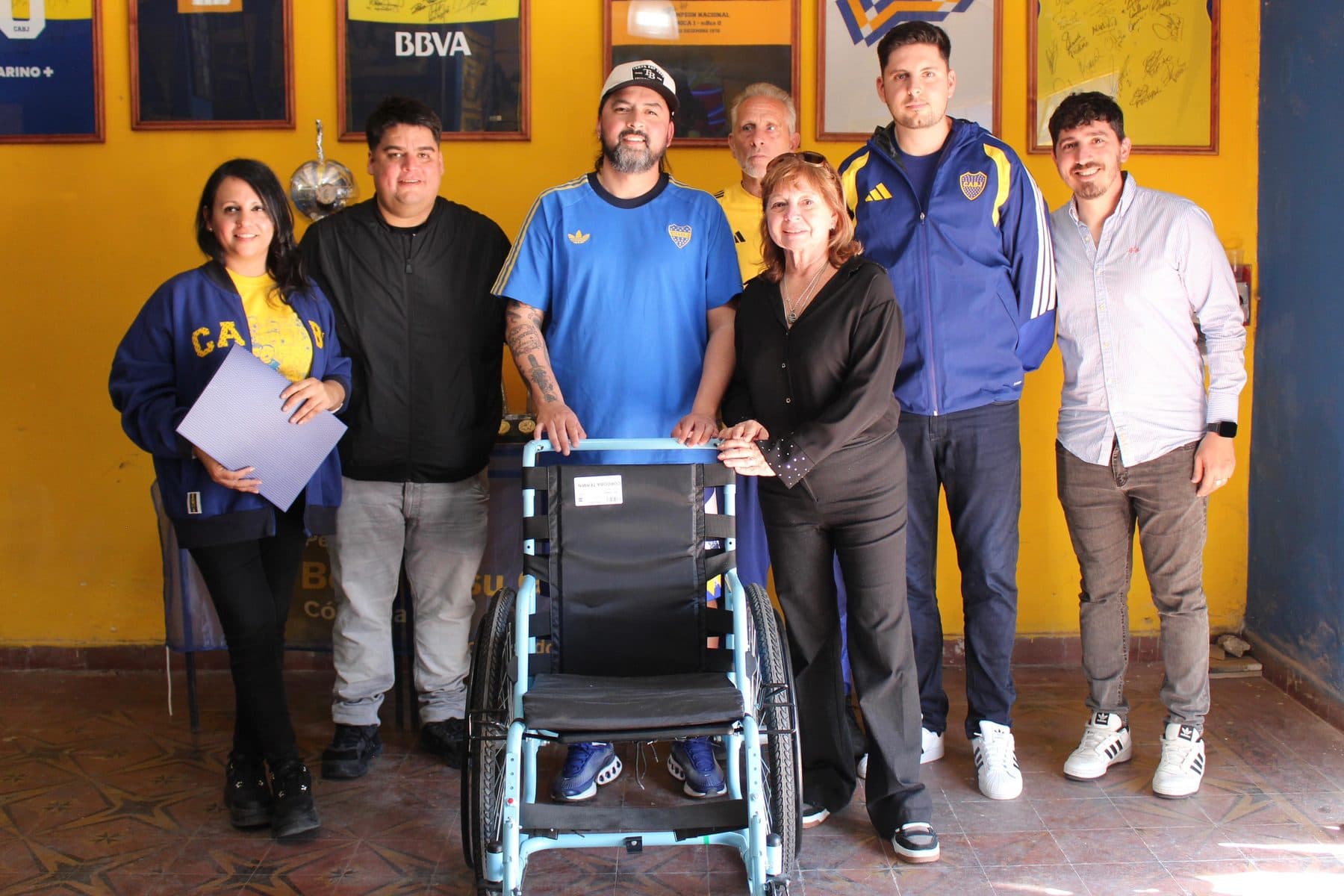El equipo de la Fundación y referentes de las peñas posando junto a una de las sillas de ruedas, en el salón de la peña, con las camisetas enmarcadas en la pared